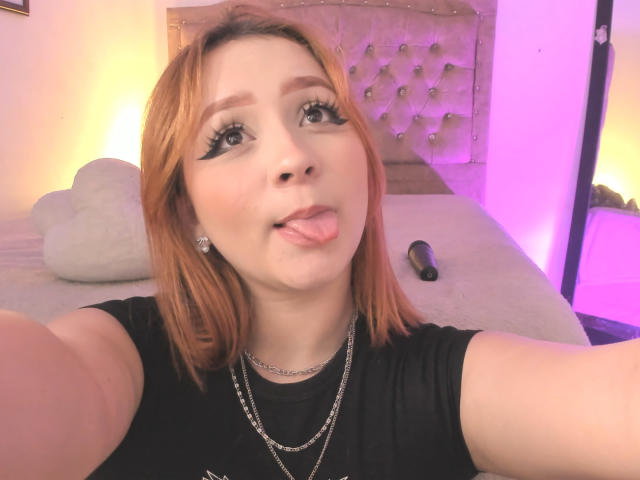 DarkCourtney with orange hair, black eyeliner, and tongue out, wearing a black shirt and necklace, in a bedroom with pink lighting, holding a black object.