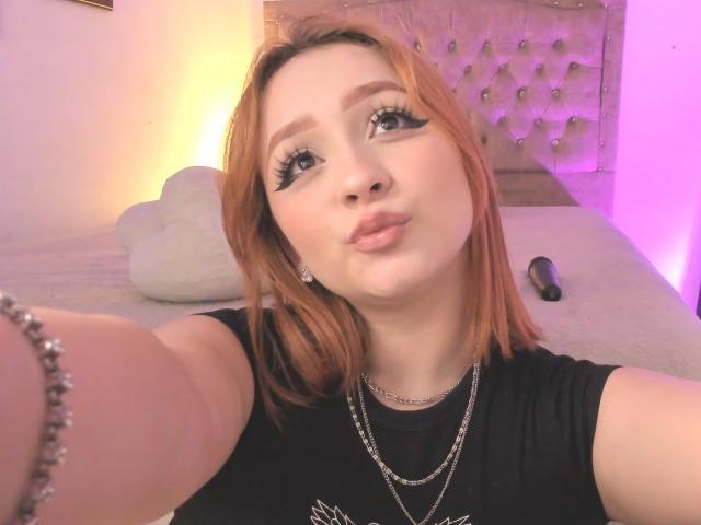 DarkCourtney with orange hair, black eyeliner, and pink lipstick, pouting, wearing a black shirt, necklace, and bracelet, in a bedroom with purple lighting. Her tits are slightly visible.