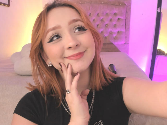 DarkCourtney has light skin, red hair, and thick black eyeliner. She's smiling with a hand near her face, wearing a black shirt and a ring. Pink lighting.