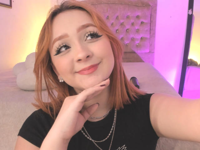 DarkCourtney with shoulder-length orange hair, black eyeliner, and pink lipstick, smiling at the camera, hand on cheek, wearing a black shirt and necklace, in a softly lit room.