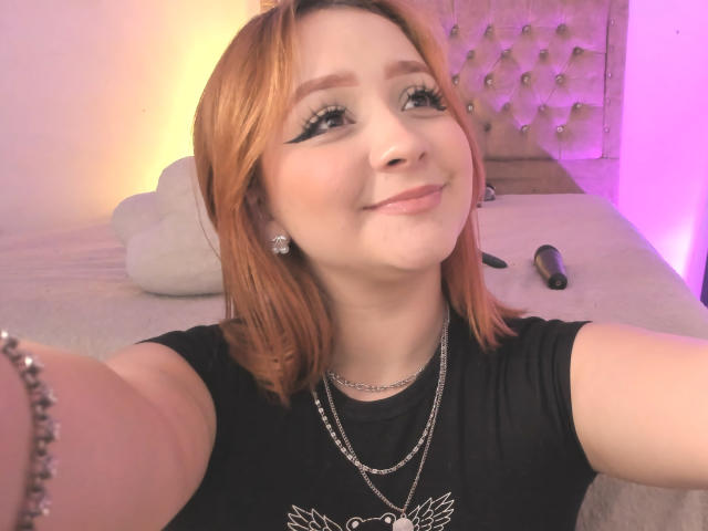 DarkCourtney with red hair, black shirt, and silver necklace, smiling at the camera, holding a selfie. Her tits are small, she's wearing a chain bracelet. Vibrant pink and yellow lights.