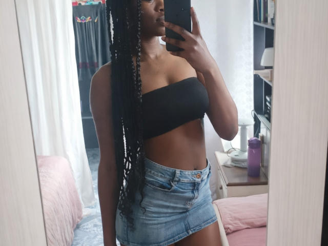 Nahomyyy with dark skin, long braided hair, black strapless top, showing her boobs, and blue denim skirt, taking a mirror selfie, holding a phone, in a bedroom.