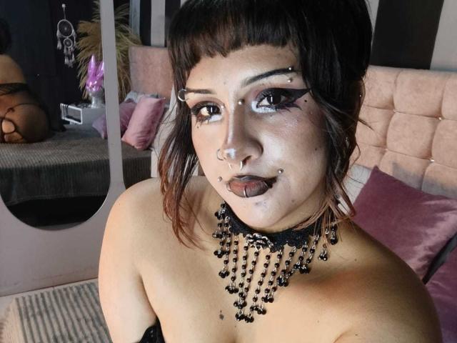 DanniLuke with dark hair, piercings, black eyeliner, tongue ring, black choker with beads, and exposed boobs, looking at camera. Reflection shows her ass in black lingerie.