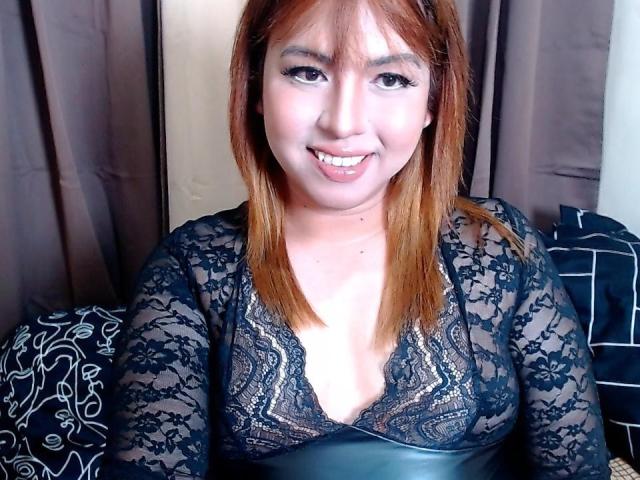 TsDominantMichaela with light brown hair, smiling, showing teeth, wearing a black lace top with deep V, exposing cleavage, sitting on a bed with patterned pillows.