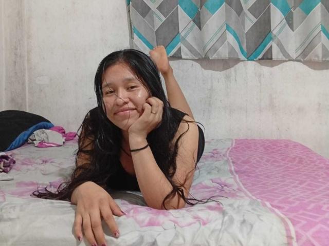 Guess69 with long black hair, lying on a bed, legs up, smiling, black sleeveless top, right hand on chin, left hand on bed, pink and white patterned blanket.