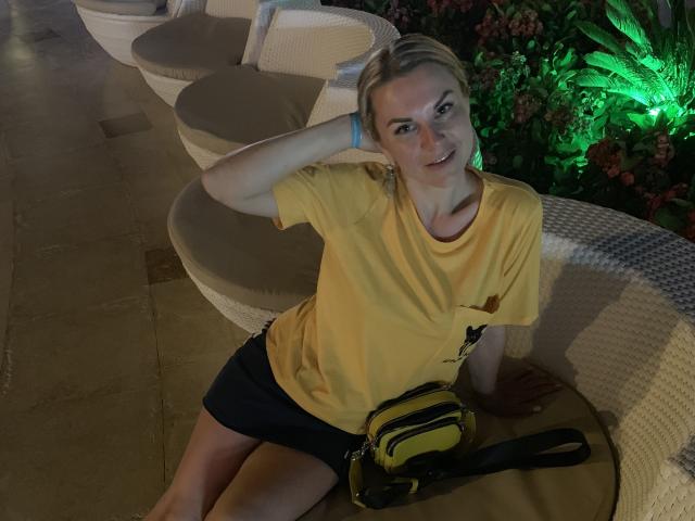 BelleLisaG with blonde hair, yellow shirt, black shorts, and yellow bag, sitting on a wicker chair, hand in hair, smiling, green plant light in background, night setting.
