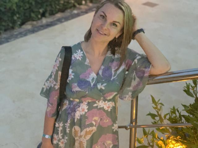 BelleLisaG con cabello castaño claro, sonriendo, vestida con un vestido floral verde, con tirantes negros y un reloj de pulsera, apoyada en una barandilla de metal, planta verde en la esquina inferior derecha..
