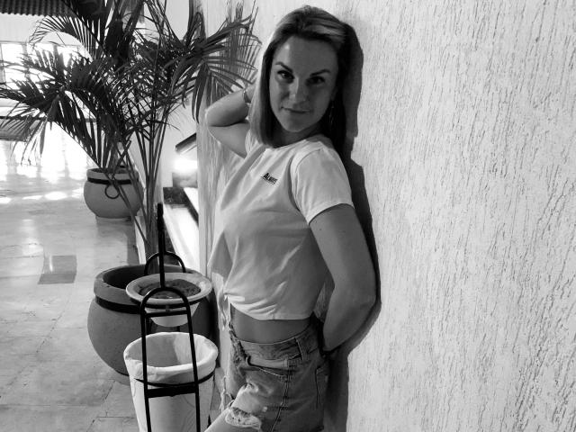 BelleLisaG with shoulder-length hair, white tee, and ripped jeans, leaning against a textured wall, hand in hair, small tits, subtle smile, black-and-white, potted plant on left.
