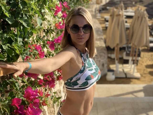 BelleLisaG with blonde hair, wearing a green leaf-patterned bikini top, dark sunglasses, and a blue bracelet, standing against a pink bougainvillea wall, showing her toned abs.