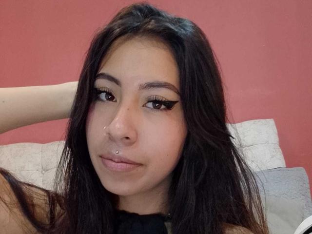 LinaSpearce with long dark hair, brown eyes, winged eyeliner, nose piercing, and lip piercing, hand in hair, looking at camera, red wall in background, white pillow behind.