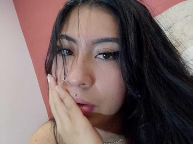 LinaSpearce a de longs cheveux noirs, un piercing au nez, et une main qui recouvre légèrement ses lèvres rouges. Elle a des yeux foncés, un maquillage discret, et une expression neutre..