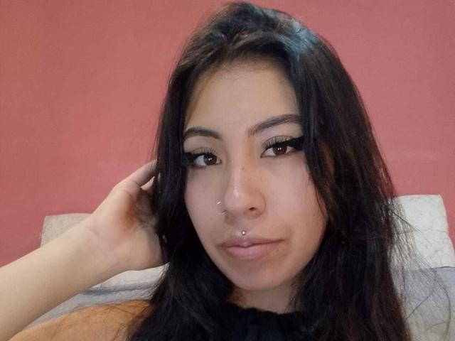 LinaSpearce ha lunghi capelli neri, un piercing al naso e un piercing al setto, con un eyeliner nero intenso e un sorriso appena accennato, con una mano appoggiata sulla testa..