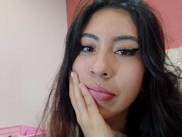 LinaSpearce a de longs cheveux noirs, des yeux marron et un piercing au nez. Elle repose sa main sur ses lèvres roses, et regarde légèrement sur le côté avec un sourire discret..