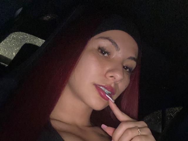 ValeryHarllow with dark red hair, wearing a black hijab, licking her glossy pink lipstick with her finger, looking seductive, wearing a ring, in a dimly lit car.