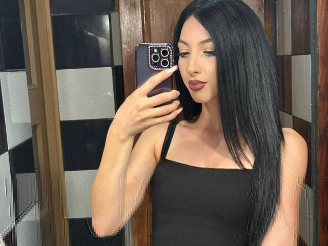SucreVixen with long black hair, wearing a black tank top, taking a selfie in a mirror, holding a phone with a purple case, showing pink nails, and a slight smile.