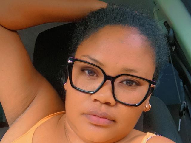 Mevah with medium brown skin, black glasses, and short curly hair, raising an arm, wearing a yellow top, showing her face and a neutral expression.