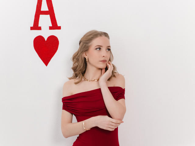 EvaDaly, a blonde woman with wavy hair, wears an off-shoulder red dress, a gold necklace, and a bracelet. She stands against a white wall, looking to the right with her hand touching her cheek. A red "A" and heart are on the wall to her left. Her expression is calm and contemplative. The red dress contrasts with her fair skin and the white background. The composition is simple and focuses on her. The lighting is even, highlighting her features and the red elements.