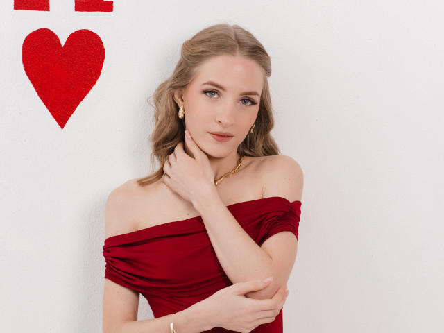 EvaDaly stands against a white wall, wearing a red off-shoulder dress that clings to her form. Her right hand touches her neck, her left arm crosses her chest. She has light skin, blue eyes, and wavy blonde hair. A gold necklace and small earrings are visible. A red heart is affixed to the upper left. Her expression is calm, slightly smiling. The image is sharp, with soft lighting.