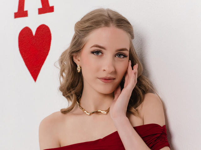 EvaDaly has blonde, wavy hair and blue eyes, wearing a red off-shoulder top and gold chain necklace. She touches her cheek with her right hand, showing a slight smile. She has gold earrings and light makeup. The white wall behind her has a red heart and "I" in the upper left. Her skin is fair, and her expression is calm and confident. The image is sharp and well-lit.