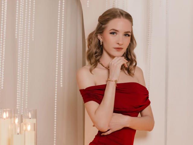 Eva Daly, met golvend blond haar, draagt een rode jurk met een open schouder, gouden oorbellen en een armband, en poseert met haar hand in de buurt van haar kin, kijkend naar de camera, in zacht, warm licht..