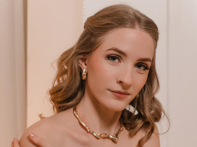 EvaDaly is a young woman with wavy, light brown hair and fair skin. She has blue eyes, subtle makeup, and wears gold earrings and a chunky gold necklace. Her expression is calm and slightly curious. The lighting is soft, highlighting her smooth skin. The close-up portrait focuses on her upper torso, with her right hand partially visible, resting on her left shoulder. The background is blurred, keeping the focus on EvaDaly.