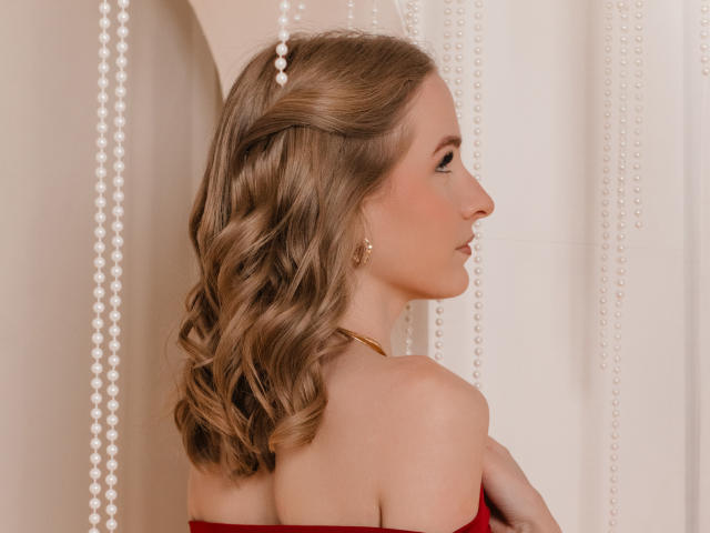 EvaDaly in a side profile, facing right. She has wavy, light brown hair with a small braid on the left. She wears a red strapless top, gold earrings, and a gold necklace. Pearl strings hang vertically in the background. Her skin is fair, and she has a small nose. The composition is clean with a soft, warm color palette. The lighting is even, highlighting her profile and hair texture.