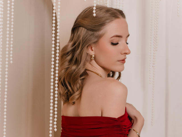 EvaDaly with wavy blonde hair, red strapless top, gold earrings, and pearl beads, looking down, holding her tits, soft light, elegant, sexy, graceful.