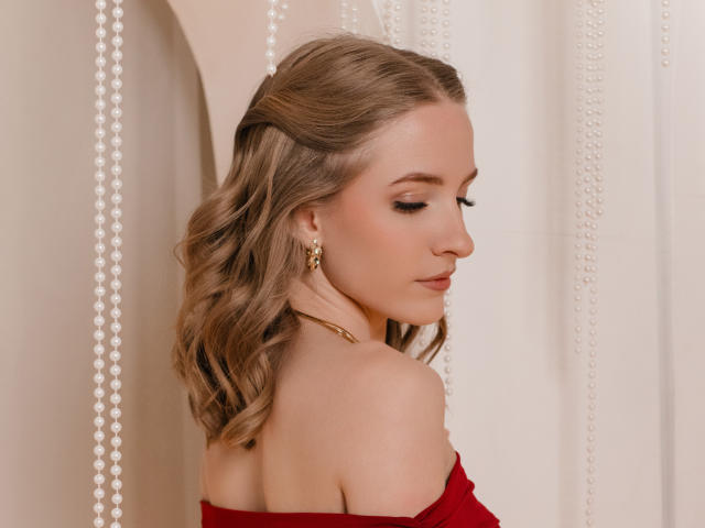 EvaDaly with wavy blonde hair, red off-shoulder top, gold earrings, and closed eyes, showing her bare shoulders and neck. Beads hang in the background.