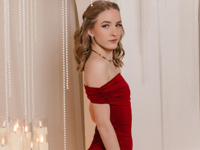 EvaDaly stands in profile, wearing a red, off-the-shoulder dress that ruches at the chest. She has wavy, light brown hair, a gold chain necklace, and subtle makeup. Pearl strings hang in the background. On the left, several lit candles are in clear holders. EvaDaly's skin is fair, and she has a slight, confident smile. The image has a soft, elegant aesthetic.