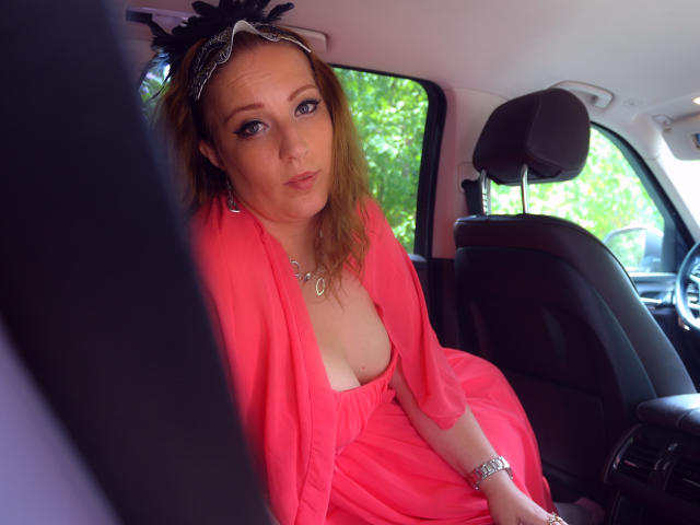 VanessaSquirtt sits in a car, wearing a bright pink, deep V-neck dress with a sheer, flowing fabric. She has fair skin, blue eyes, and shoulder-length blonde hair. A black lace headpiece with feathery adornments sits on her head. She wears a silver necklace and a bracelet on her right wrist. The car's black seat and headrest are visible, with green foliage outside the window. Her expression is neutral, slightly curious. The photo is taken from a slightly low angle.