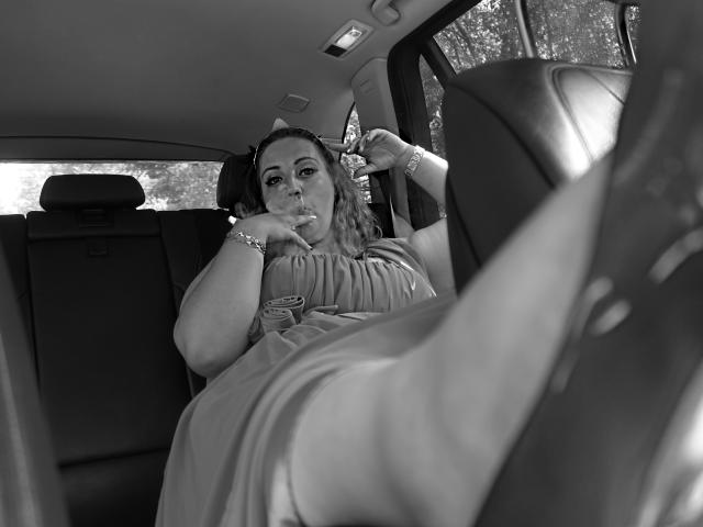 Black and white photo of VanessaSquirtt in a car. She's wearing a dress, sitting in the back seat, and smoking a cigarette with a relaxed expression. Her right arm rests on the car seat, and her left arm is raised, adjusting her hair. Her legs are extended towards the front, and a car seat is blurred in the foreground. Natural light filters through the car windows. The image has a candid, casual vibe with a focus on VanessaSquirtt's relaxed posture and the car's interior.