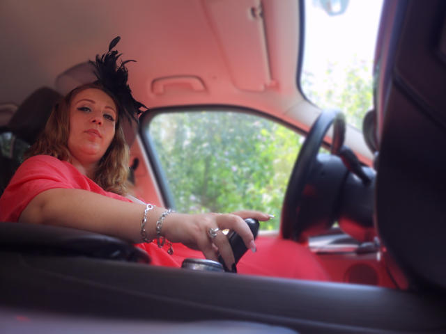 VanessaSquirtt with wavy blonde hair, wearing a black feathered hat and red dress, holding the car's steering wheel, looking forward, with a silver bracelet and rings.