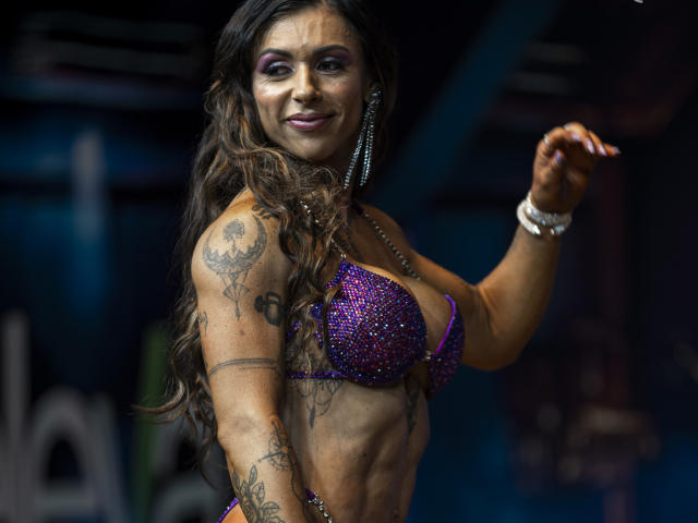 PriBttencourt with long brown curly hair, purple sparkly bra, tattoos on arms and shoulder, smiling, right arm raised, wearing white bracelet, purple eyeshadow, dark makeup, muscular, confident.