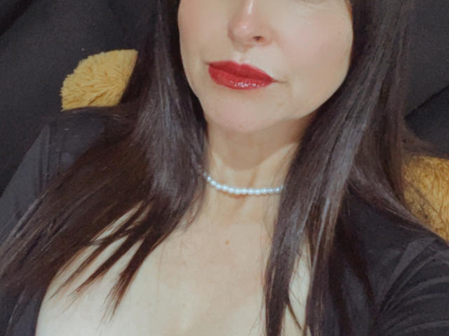 Close-up of KynkyFetish, a middle-aged woman with fair skin and red lipstick. She has straight, dark brown hair, wearing a black cardigan that reveals a modest cleavage. A white pearl necklace is visible around her neck. The image focuses on her face and upper chest, with a yellow, fuzzy pillow partially in the background. The photograph has a slightly blurry texture.