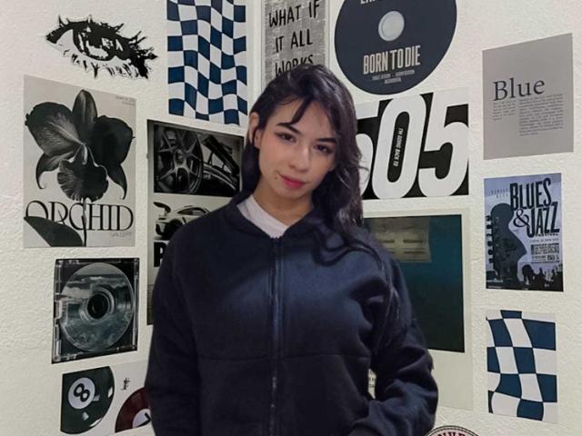KatherinJohns has dark hair, light skin, and wears a black zip-up hoodie. She's standing in front of a wall with various black and white posters.