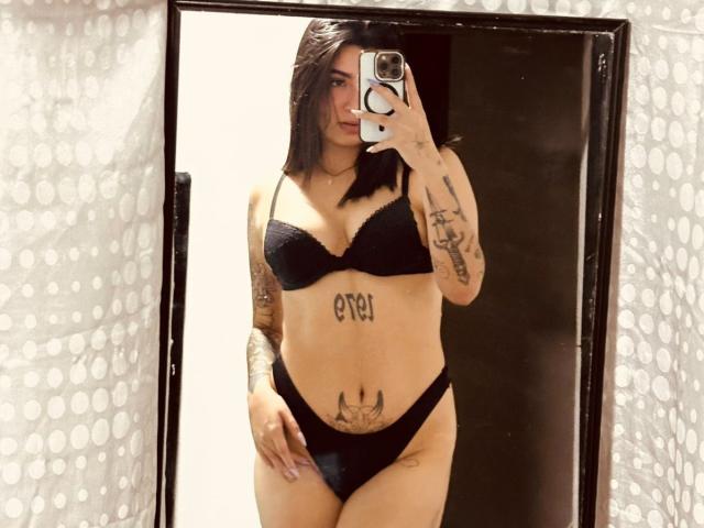 JessiParker with dark hair, black bra, and black panties, standing in front of a mirror, showing tattoos on her chest, stomach, and arm, holding a phone.