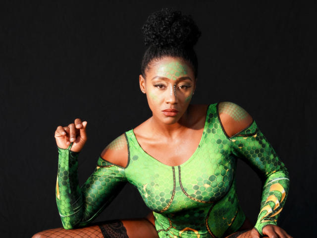 NahomyHawk is a dark-skinned woman with a high, curly black afro. She's wearing a green, long-sleeve bodysuit with hexagonal patterns and cut-out shoulders. Her face is painted with green and yellow swirls. She's seated, right hand raised, with black fishnet stockings. The bodysuit has a zipper down the center and a green belt. The composition is centered and uses a black background. Her expression is serious and focused.