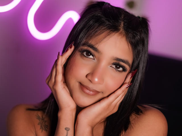 Scarlesth with dark hair, brown eyes, and red nails, hands on cheeks, smiling softly. She has tattoos on her right shoulder, pink neon light in background.