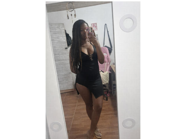 Mirror selfie of Nahomyyy, standing sideways, wearing a tight black leather mini dress with a side slit. Nahomyyy has long, straight, dark brown hair, wearing glasses, and holding a phone in her right hand. Her right arm is slightly raised, showing off her cleavage and toned legs. She has a medium to dark skin tone. The room has a wooden floor, a pink gaming chair, and a white door with a dreamcatcher. Nahomyyy's phone case is pink with a floral pattern. Her footwear is strappy sandals.