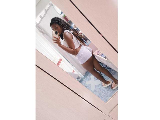 Nahomyyy stands in front of a mirror, taking a selfie. She's wearing a white lace bralette and matching lace shorts, with long, styled braids. She's looking over her shoulder, showing off her curvy, toned figure. She's wearing white high-heels. The mirror is in a wooden closet, reflecting a patterned blue rug. The image has a bright, natural light. Nahomyyy's skin is dark, and she has a confident, sexy pose.