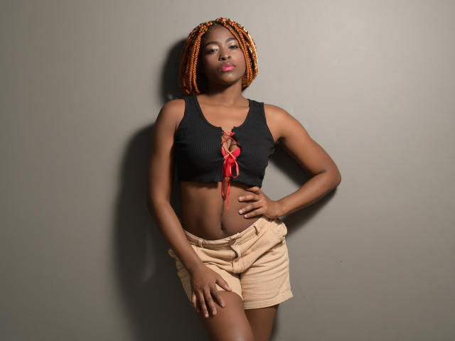 NicoleHills has orange braided hair, dark skin, small tits in a black lace-up crop top, and beige shorts, standing with hands on hips, looking confident.