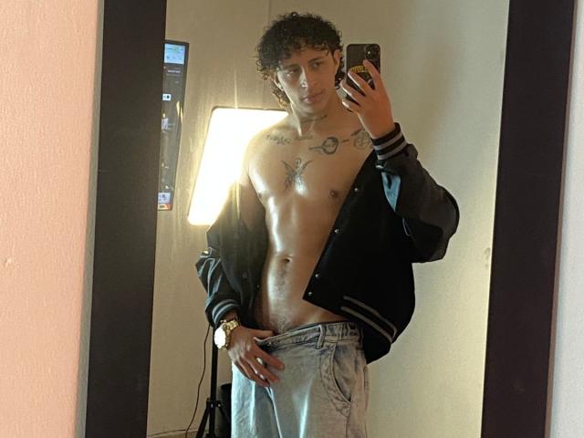 Jay Taylor, com cabelo cacheado, vestindo uma jaqueta preta aberta, exibindo um peito musculoso e tatuado e abdômen plano, segurando um telefone, em pé em frente a um espelho, vestindo calças jeans largas..