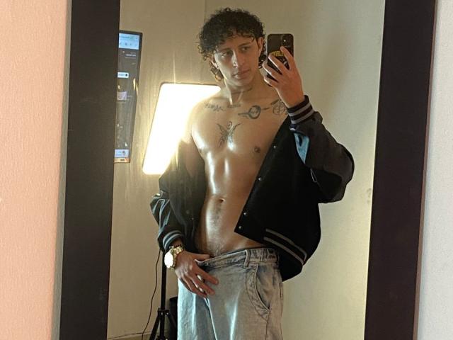JayTaylor, senza camicia, indossa una giacca nera, tiene in mano un telefono, sta scattando un selfie allo specchio, mostrando un petto muscoloso e tonico con tatuaggi, e un leggero rigonfiamento nei pantaloni grigi..