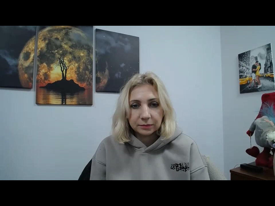 Blond Lolitti with shoulder-length hair, wearing a grey hoodie, sits in front of three moonlit paintings. Red lamp with stuffed animal on right. Slightly smiling, serious expression.