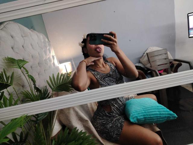 AriaVega with medium brown skin and black braids, wearing a snake-pattern dress, taking a mirror selfie, tongue out, sitting on a chair with a teal pillow.
