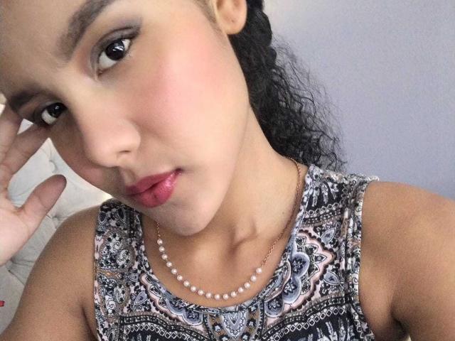 AriaVega, con i capelli neri ricci, indossa un abito a fantasia e una collana di perle, ha un leggero rossore sulle guance e guarda la fotocamera con le labbra rosa leggermente dischiuse..