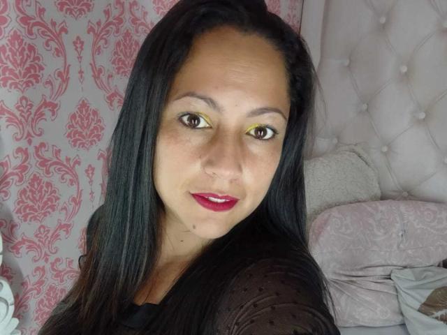 ScarlettBarness with yellow eyes, red lipstick, and black hair, wearing a sheer black top, looking at the camera with a slight smile. Her tits are slightly visible.