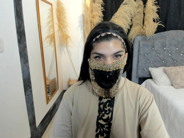 AniisaAli with long black hair wears a gold headpiece and a black mask adorned with gold chains and intricate patterns. She has brown eyes and wears a beige robe with a black patterned shirt underneath. Her head is tilted slightly, and she is in a bedroom with a gray tufted bed and a mirror on the left. Pampas grass is in the background. The image has a casual, selfie-like quality.