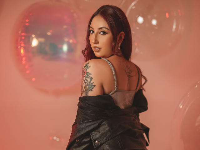 EmiilyWatson with long red hair, looking back, wearing a black leather jacket off her shoulders, exposing her back tattoo and gray tank top, showing her small, cute ass.