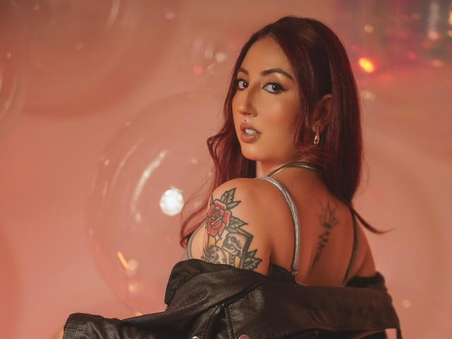 EmiilyWatson with red hair, looking back, showing tattooed shoulder with rose, wearing off-shoulder top, black jacket, and earrings, in soft pink light. Boobs visible, ass not.